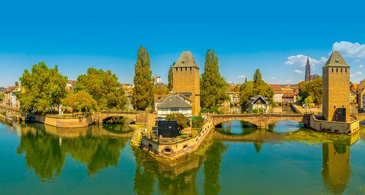 Vue panoramique des ponts couverts historiques et des tours de Strasbourg se reflétant dans les eaux calmes de la rivière sous un ciel bleu dégagé.