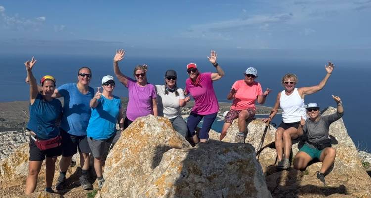 Neuf randonneurs posant joyeusement sur un sommet rocheux avec une vaste mer et des montagnes côtières sous un ciel bleu dégagé