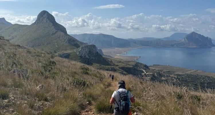 Des randonneurs marchant sur un sentier herbeux vers des falaises côtières spectaculaires et une baie turquoise sous des nuages épars
