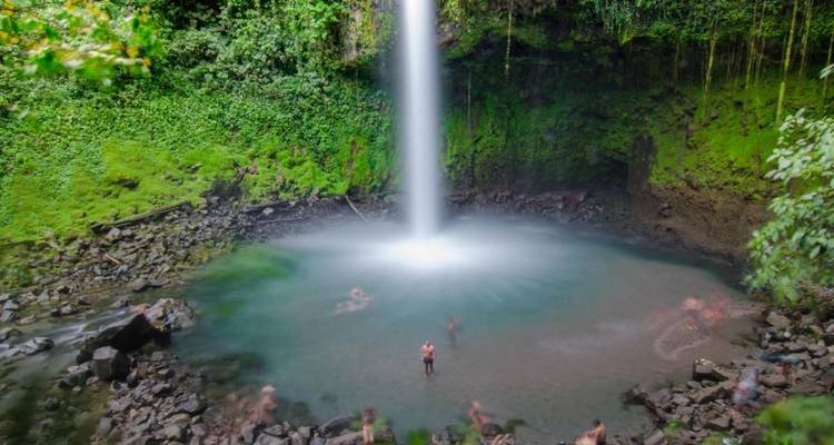 Prise de vue en pose longue de nageurs profitant d'un bassin de plongée sous une haute cascade de jungle entourée de falaises moussues.