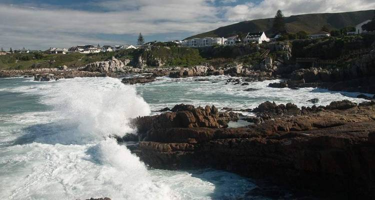 Vagues s'écrasant contre des rochers escarpés près d'un village côtier avec des montagnes en arrière-plan.