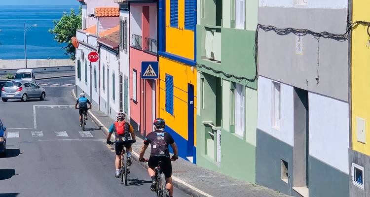 Mountainbiker fahren an leuchtend bemalten Häusern mit Blick auf den Atlantik auf der Insel São Miguel vorbei.