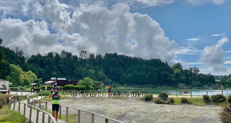 Radfahrer pausieren neben einem ruhigen Kratersee, der von üppigen Hügeln und dramatischen Wolken umrahmt wird.