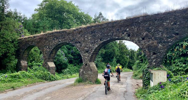 Radfahrer fahren unter einem alten Steinaquädukt hindurch, umgeben von dichter grüner Vegetation.