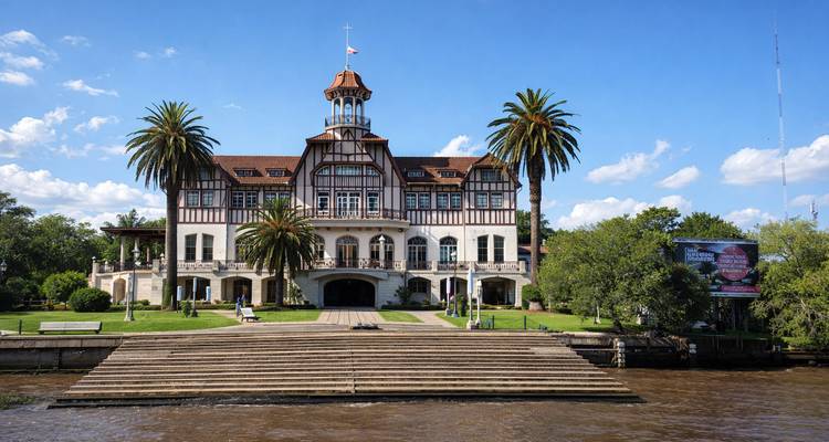 Club élégant de style colonial au bord de la rivière, flanqué de hauts palmiers, vu à travers une large étendue d'eau brune sous un ciel bleu clair.