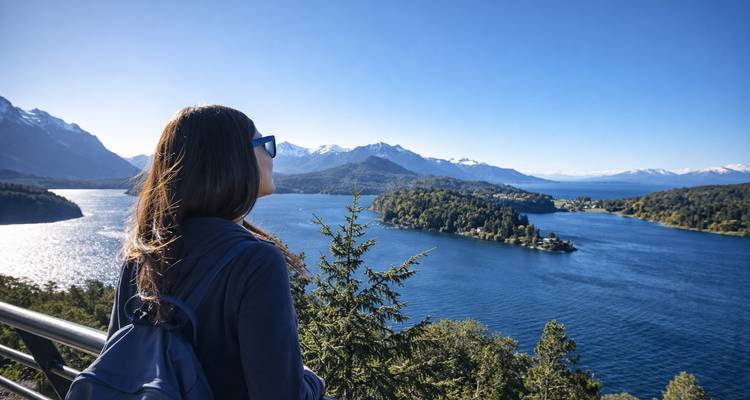 Femme avec sac à dos admirant un lac bleu profond parsemé d'îles boisées et de pics andins enneigés au-delà.