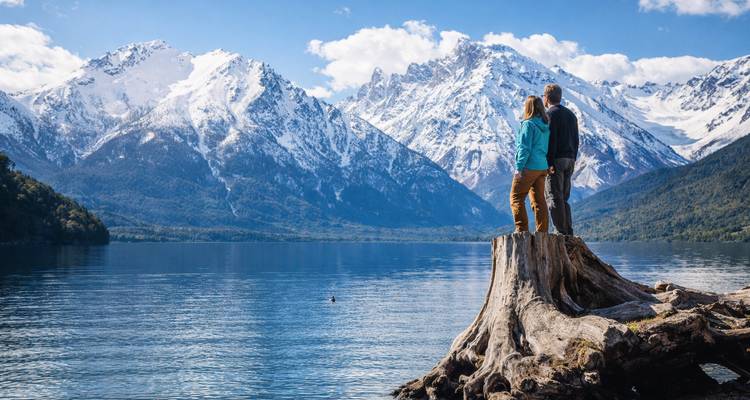 Couple debout sur une énorme souche de tronc contemplant un lac alpin calme avec en arrière-plan des montagnes enneigées spectaculaires.
