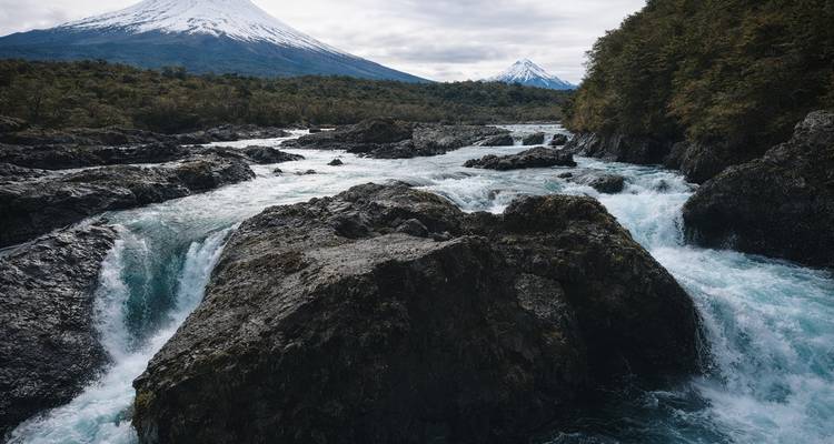 Puissante rivière turquoise cascadant sur de la roche volcanique avec un volcan conique enneigé s'élevant au loin.