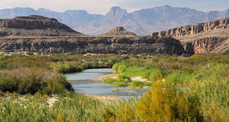 Une rivière serpentine bordée de roseaux traverse un terrain rocheux aride dominé par des montagnes désertiques escarpées.