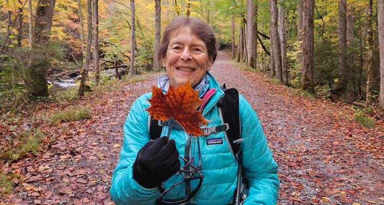 Un randonneur souriant dans une veste turquoise tient fièrement une feuille d'érable brillante sur un sentier d'automne coloré.