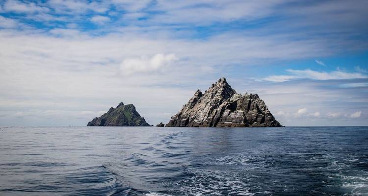 The jagged twin islands of Skellig Michael rise sharply from the Atlantic, viewed from a boat trailing wake.