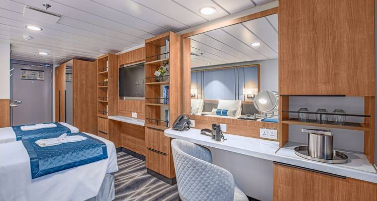 Spacious modern cruise cabin with twin beds, TV, desk, and neutral blue accents.
