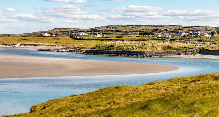 A winding tidal inlet cuts through sandy flats toward a small rural Irish village under sunny skies.