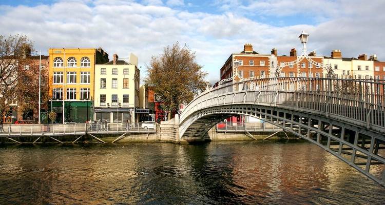 A classic cast-iron pedestrian bridge spans the River Liffey with colorful Georgian buildings lining the opposite embankment under a bright sky.