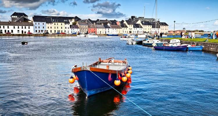 Ein leuchtend blaues Fischerboot mit Bojenfendern schwimmt in einem ruhigen Hafen vor einer Reihe pastellfarbener Stadthäuser an einem sonnigen Tag.
