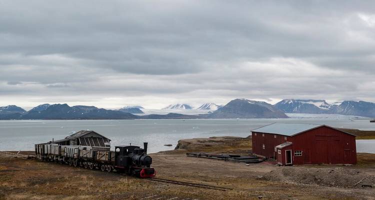 Een oude locomotief en rode schuur rusten op de Arctische toendra naast ijzige fjordwateren en met sneeuw bedekte bergen onder bewolkte luchten.