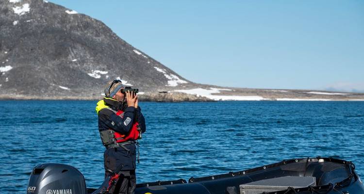Ontdekkingsreiziger in een kleine opblaasboot speurt de Arctische kust af met een verrekijker tegen een achtergrond van besneeuwde bergen.
