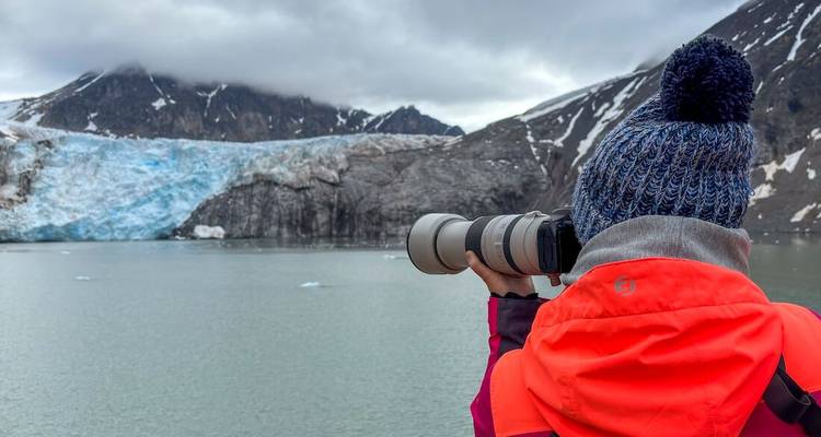 Reiziger in felgekleurde jas fotografeert een blauwe gletsjer over een kalme Arctische fjord met een telelens camera.