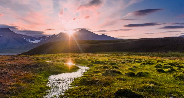 Low Arctic sunlight bursts over jagged snow-capped peaks, casting golden light on a mossy green valley and a winding melt-water stream.