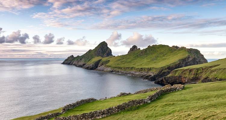 Groene kaap en zeerotsen van St. Kilda rijzen op boven kalme Atlantische wateren onder een pastelkleurige hemel.
