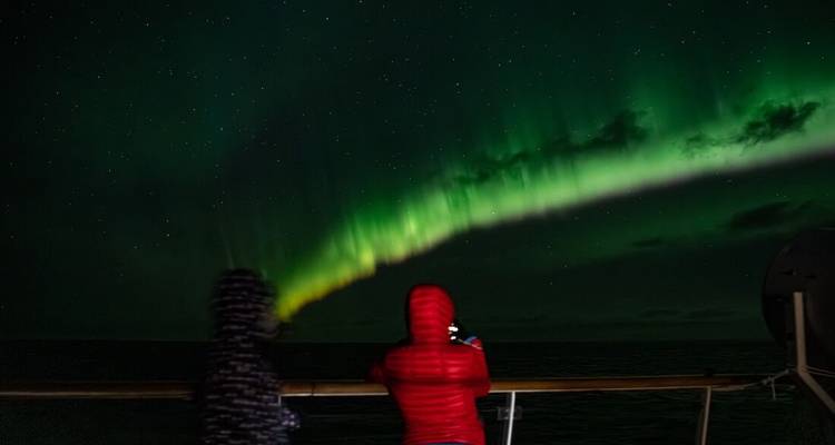 Deux passagers emmitouflés observent de vives aurores boréales vertes s'arquant à travers un ciel nocturne sombre depuis le pont d'un navire.