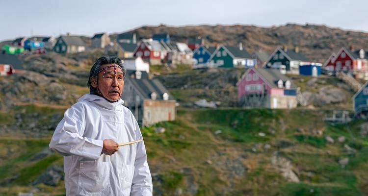 Artiste inuit en costume traditionnel tenant un tambour avec des maisons groenlandaises colorées sur une colline rocheuse en arrière-plan.