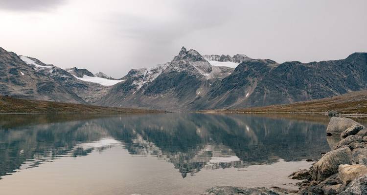 Lac alpin immobile reflétant parfaitement les pics enneigés déchiquetés et les glaciers sous un ciel tamisé.