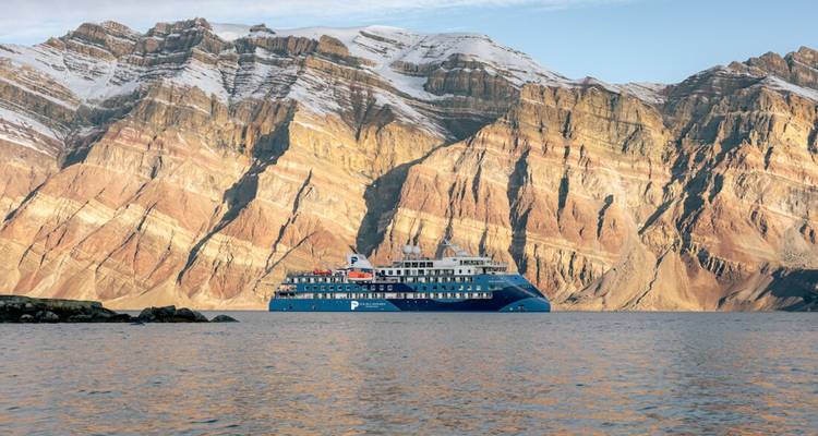 Navire d'expédition bleu navigue le long de falaises spectaculaires aux couches géologiques marquées qui s'élèvent abruptement depuis les eaux calmes de l'Arctique.
