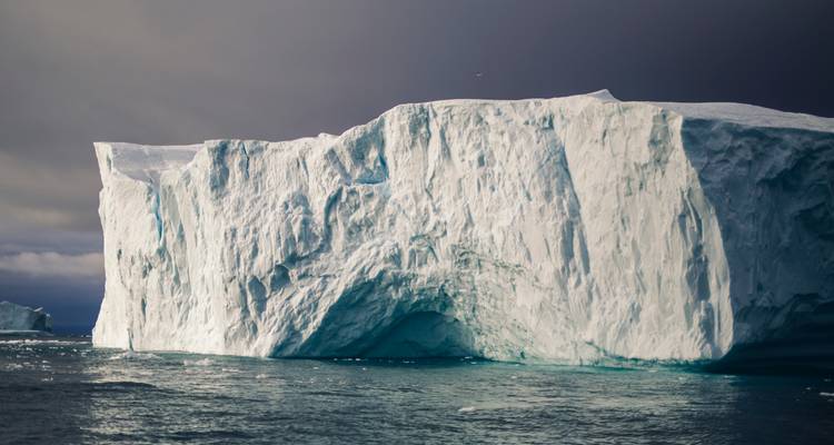Massive white iceberg with textured cliffs floating in dark Arctic waters under moody skies.