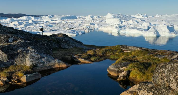 Lone traveller stands on rocky shore overlooking a field of drifting icebergs at dusk.
