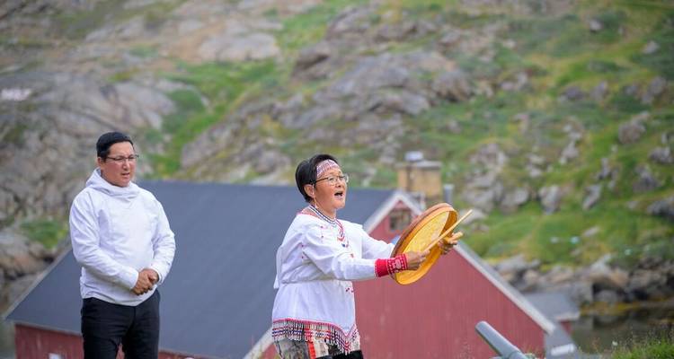 Femme en costume traditionnel groenlandais bat un tambour tandis qu'un homme regarde, maisons en bois derrière.