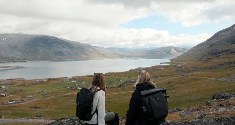 Deux randonneurs sont assis sur une pente rocheuse surplombant une vaste vallée lacustre et des habitations dispersées.
