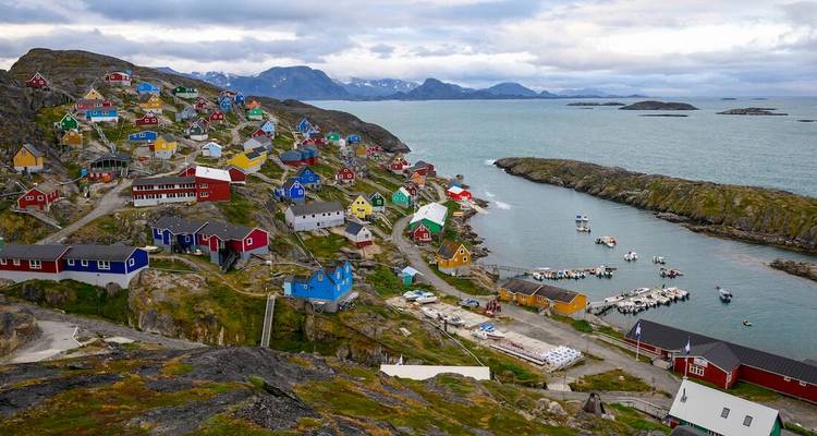 Village groenlandais à flanc de colline avec des maisons aux couleurs vives surplombant la mer sous des nuages dramatiques.