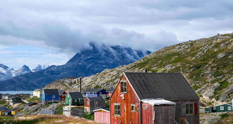 Maison en bois rouge patinée se dresse sur un terrain rocheux avec un sommet de montagne couvert de brume derrière.