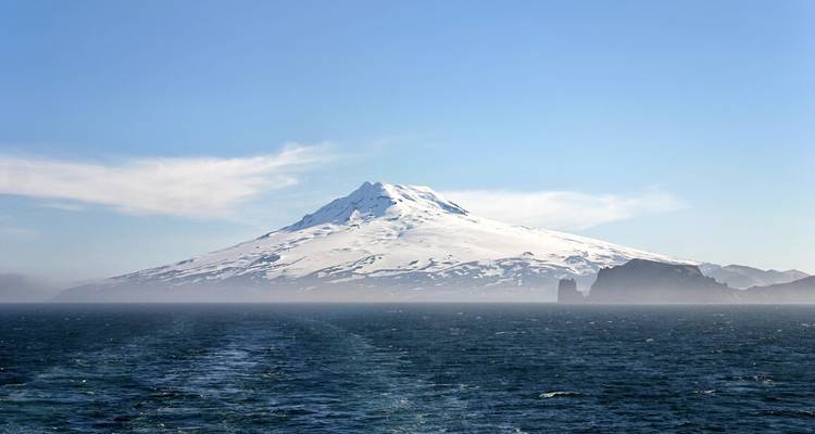 Snow-capped volcanic island peaks above a misty sea with a distant ship wake leading toward it.