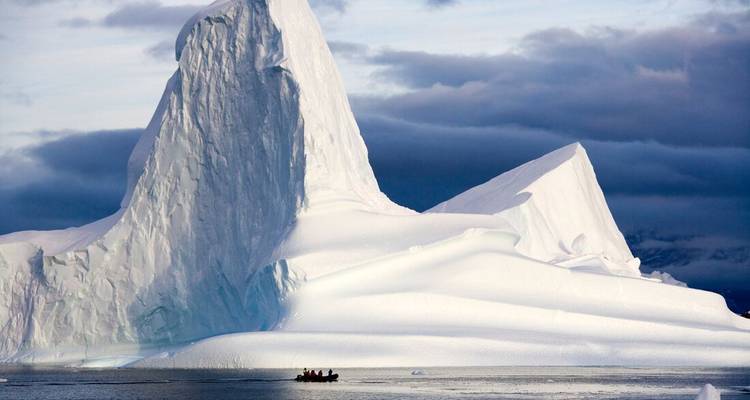 Huge sculpted iceberg towers over a tiny zodiac boat on calm polar waters under dramatic skies.