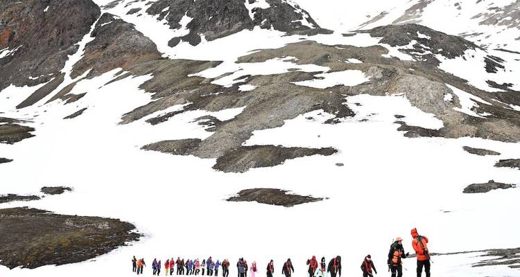 Line of bundled explorers trekking up a snow-covered Arctic hillside dotted with rocks.