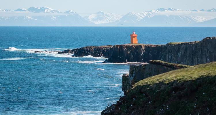 Een levendig oranje vuurtoren staat op een ruige IJslandse kaap met uitzicht op diepblauwe zeeën en verre besneeuwde bergketens.