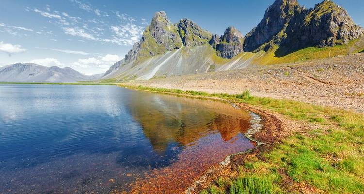 Sharp rocky peaks reflect perfectly in a still lagoon beside a stony Icelandic shoreline.