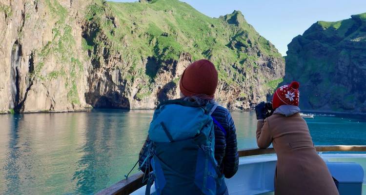 Two travellers on a boat admire towering green sea cliffs under clear skies.