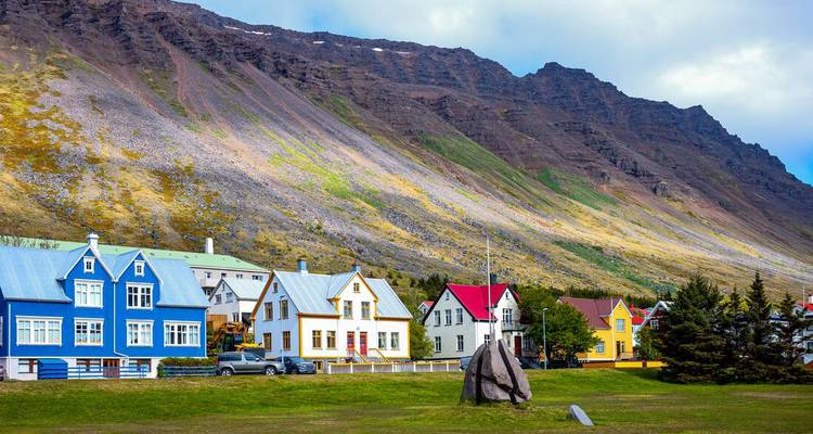 Rows of brightly painted wooden houses sit beneath a steep, striated mountain in an Icelandic fjord town.