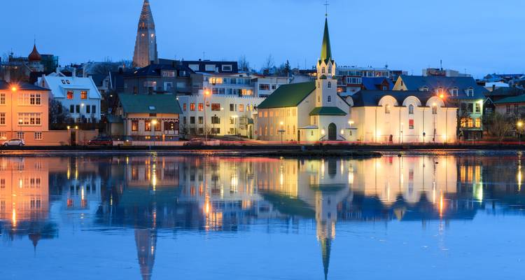 Paisaje urbano nocturno de Reikiavik reflejado en las aguas tranquilas del lago, con una iglesia de techo verde y la aguja de Hallgrímskirkja iluminada contra el crepúsculo azul profundo.