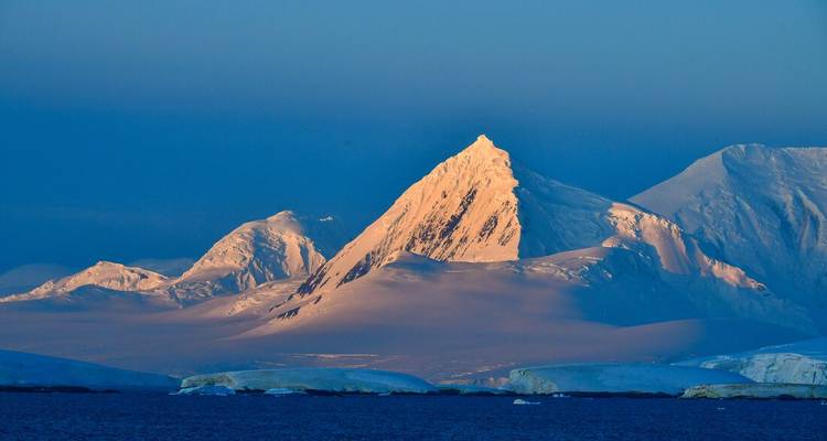 Schneebedeckter antarktischer Gipfel leuchtet golden im tiefen Sonnenlicht über tiefblauem Meer und Eisbergen.