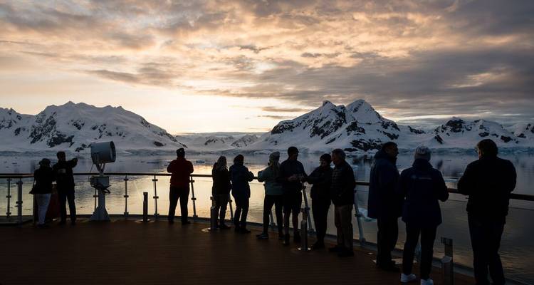 Pasajeros en silueta en la cubierta de un barco admiran las montañas antárticas cubiertas de nieve al atardecer.