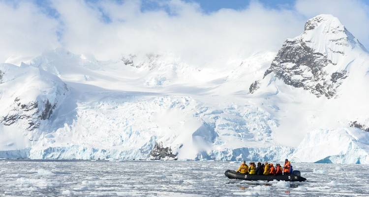 Zodiac with bundled guests floats among sea ice facing a towering white glacier wall.