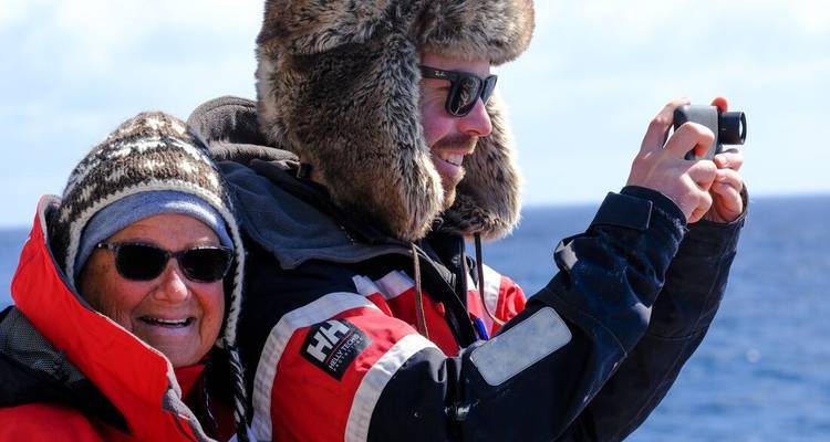 Bundled passengers on deck photograph scenery; woman smiles wearing knit hat and sunglasses.