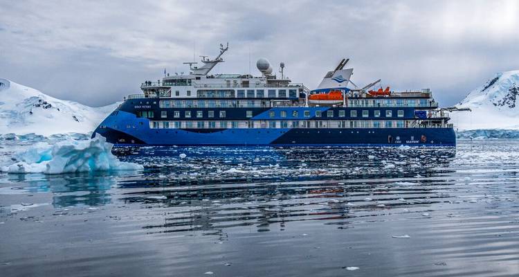 Large blue expedition cruise ship floats among brash ice against snowy Antarctic hills.