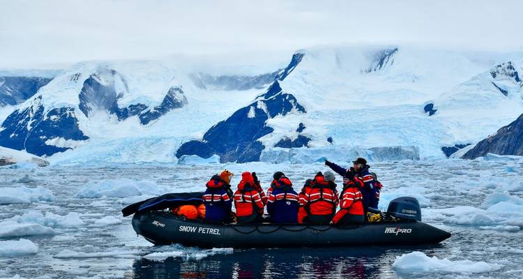Guide points from zodiac packed with guests toward vast blue-white glacier panorama.