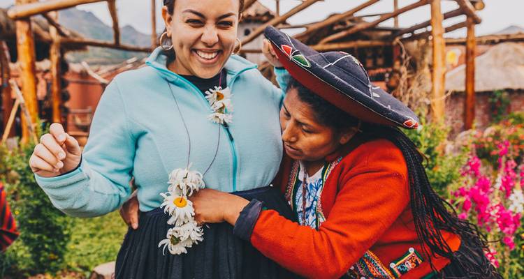 Une femme quechua locale orne de manière enjouée un voyageur qui rit avec des guirlandes de fleurs dans un cadre de village rustique.