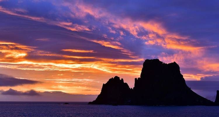 Vivid purple, orange and pink clouds blaze above a jagged sea stack silhouette at dusk.